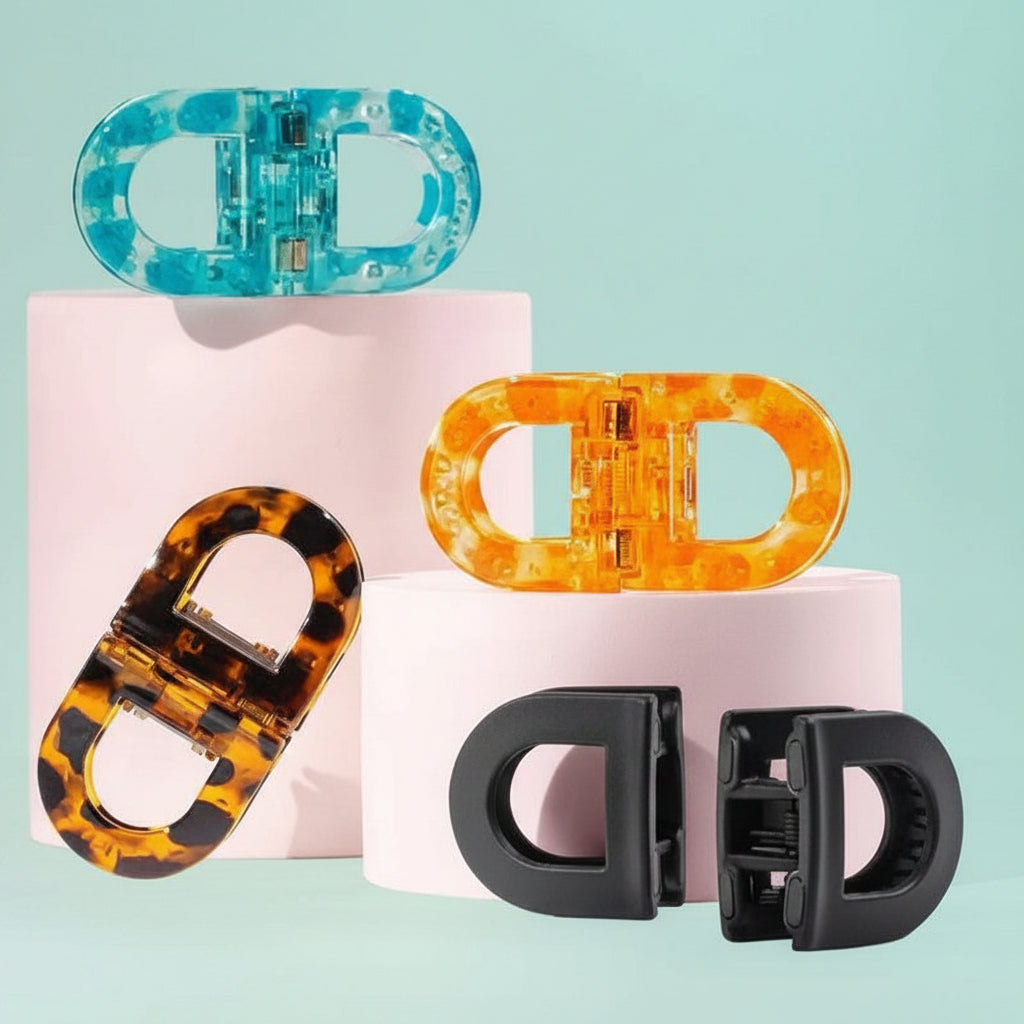 Four D-shaped hair claw clips in different colors and materials, including translucent blue, amber orange, tortoiseshell, and solid black, displayed on pink cylindrical pedestals against a light blue-green background.