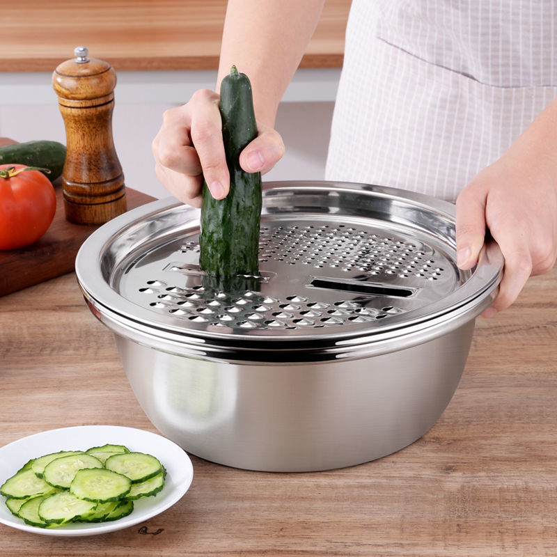 Person using a metal mandoline slicer with basin to cut a cucumber on a wooden countertop.