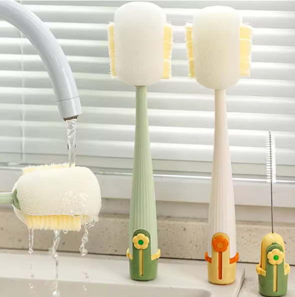 Three soft-bristle cleaning brushes with long handles stand on a bathroom or kitchen sink in front of window blinds, with running water from a faucet and one brush being washed under the stream. The handles are light green and off-white, with small decorative flower and tool accents in yellow, orange, and green.