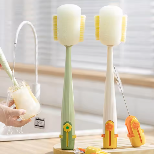 Two long-handled toothbrush-style cleaning tools with yellow bristles are shown in a bright bathroom-like setting. One is light green and one is white, both with small flower-shaped accents near the bottom.