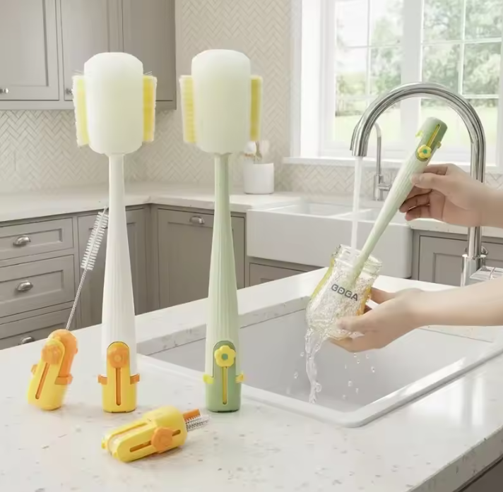 A set of Multi-functional Bottle Brush in white, mint green, and yellow, shown in a kitchen sink setting with a person's hands rinsing a glass jar. A cleaning brush and two yellow attachments are also visible on the counter.